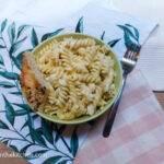 Creamy Mac n' Cheese in a green bowl, with a piece of bread on the side and a fork. Placed on two table cloths, one pink gingham and one with green leaves.