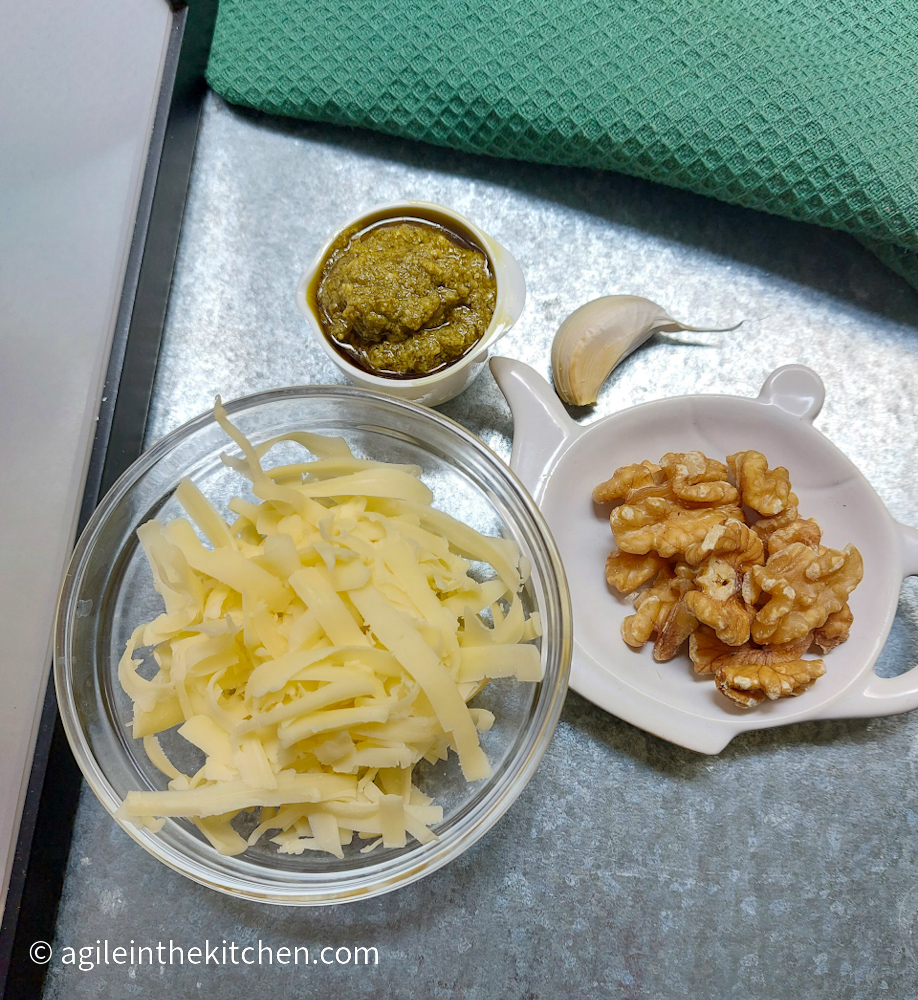 Green Mac n' Cheese ingredients laid out on a silver background: Green pesto, garlic, walnuts and shredded cheese.