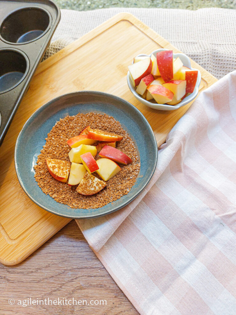 Cinnamon and sugar mixed on a blue plate, with cut up red apples being coated by the mixture. Sitting next to more cut up red apples in a bowl