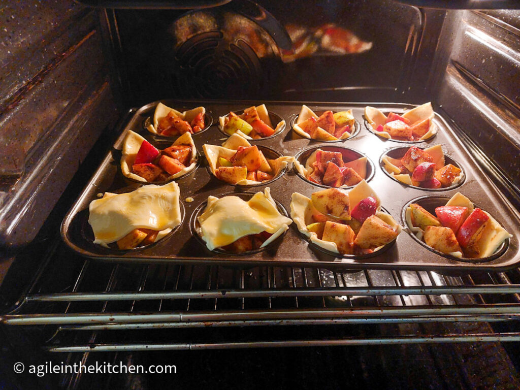 Puff pastry apple cups in the oven ready to bake. The cups are sitting in a 12 cup muffin tin. Two of the cups have a puff pastry lid.