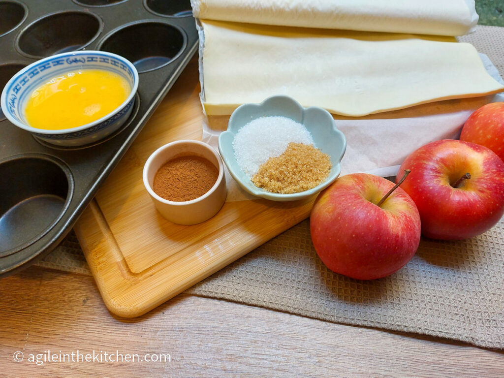 Puff pastry apple cup ingredients laid out on a background of a beige table cloth, a wooden cutting board and a muffin tin; egg wash, cinnamon, white and brown sugar, puff pastry sheet, three red apples