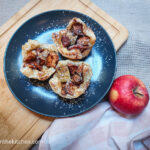 Three puff pastry apple cups straight from the oven, plated on a black plate and dusted with powdered sugar. In the background, a red apple, a pink gingham cloth and a wooden cutting board