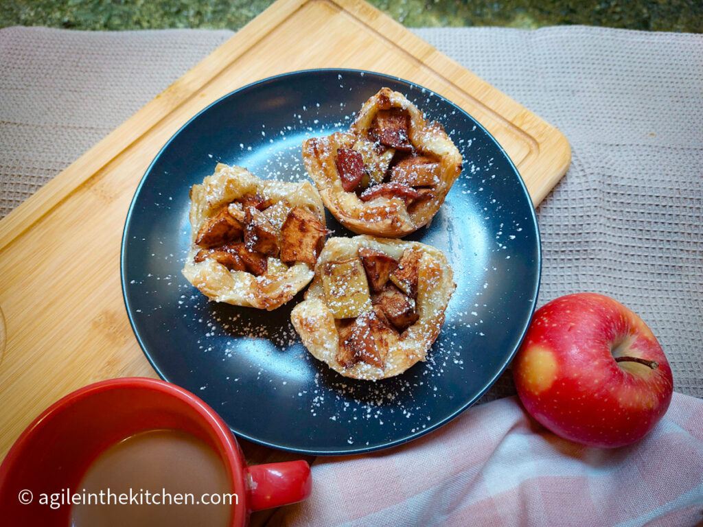 Puff pastry apple cups straight from the oven, plated on a black plate and dusted with powdered sugar. In the background, a red apple, a red cup with coffee, a pink gingham cloth and a wooden cutting board