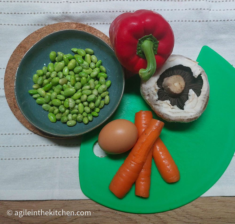 Toppings for a Ramen laid out on a white table cloth. Green edamame beans on a blue plate, a red bell pepper, a flat cap mushroom, three carrots and an egg