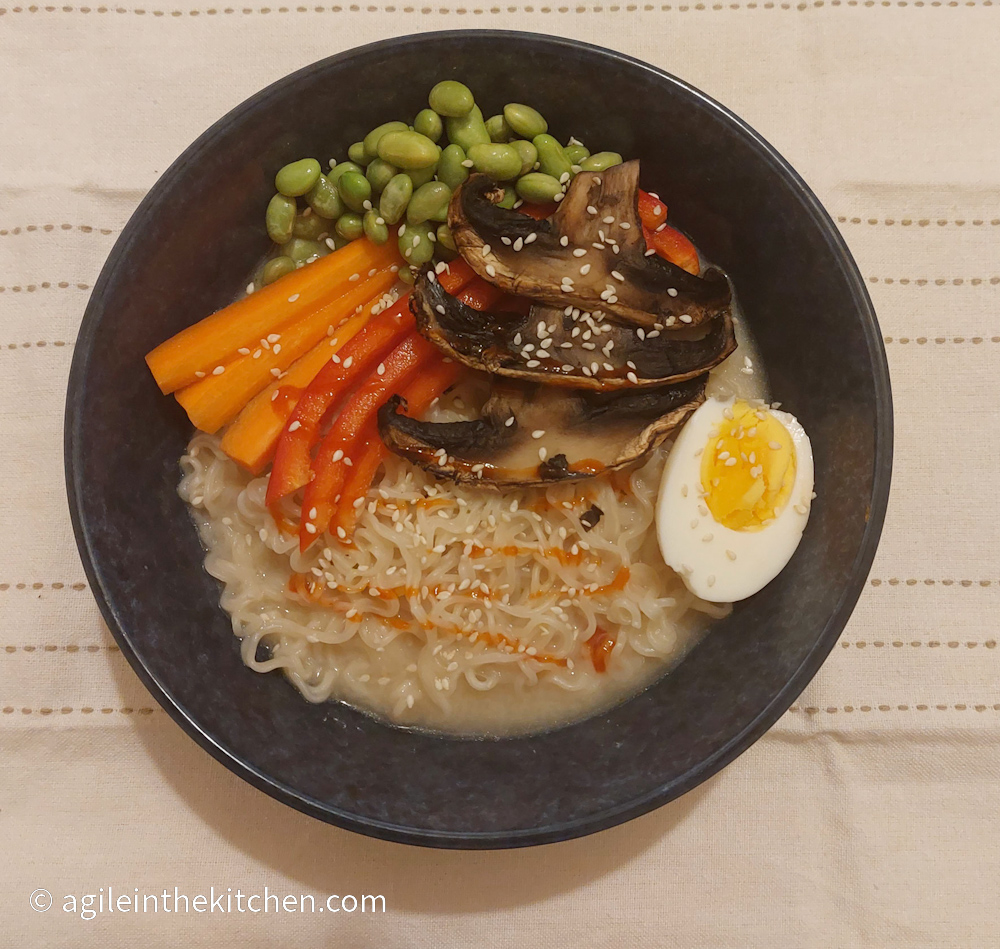 Finished Ramen laddled in to a blue bowl on a white table cloth. Ramen is topped with half a cooked egg, sliced carrots, red bell peppers and mushroom, also edamame beans and drizzled with red Shiraza.