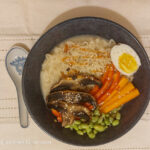 Finished Ramen laddled in to a blue bowl on a white table cloth and a spoon next to the bowl. Ramen is topped with half a cooked egg, sliced carrots, red bell peppers and mushroom, also edamame beans and drizzled with red Shiraza.