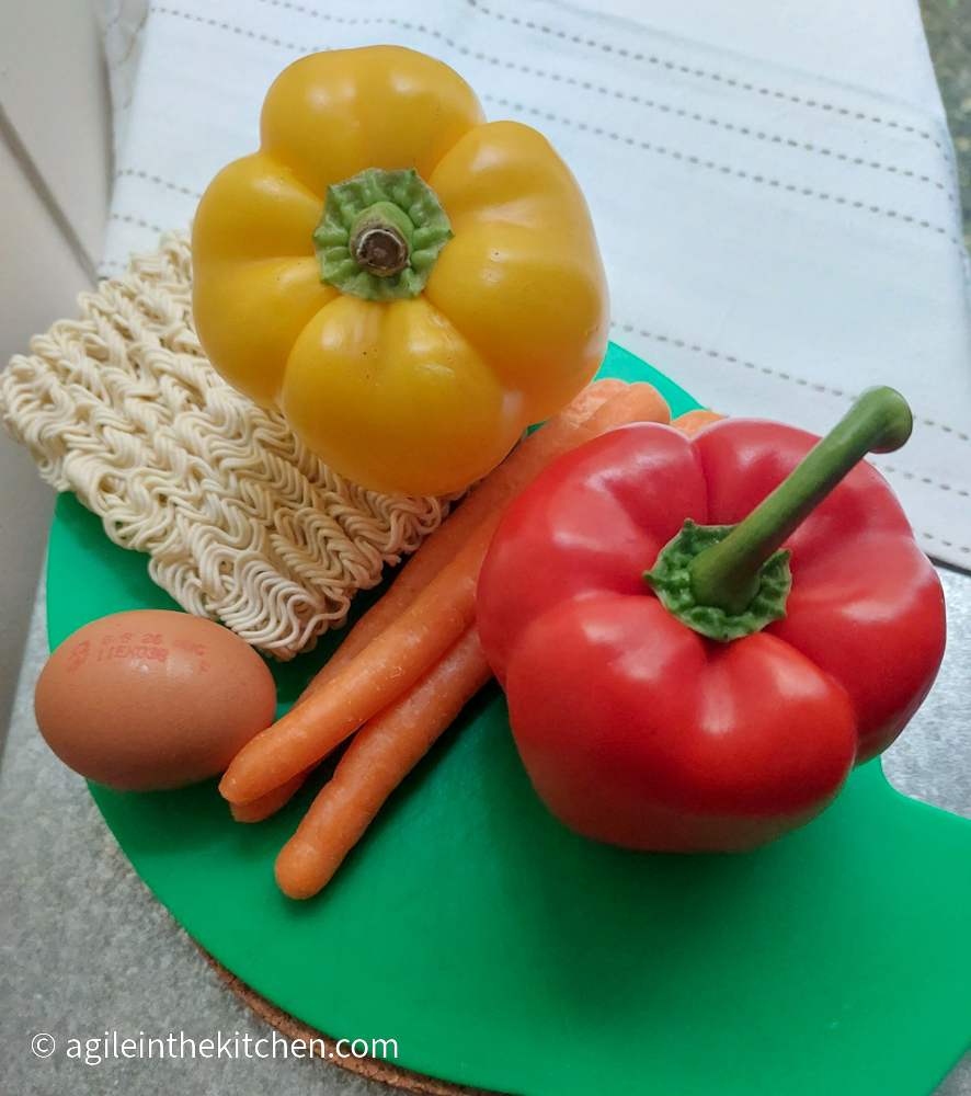 Ramen toppings laid out on a green cutting board and a white table cloth. A package of ramen, a yellow bell pepper, a red bell pepper, three carrots and an egg