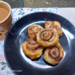 Cinnamon swirls laid on top of each other on a black plate, next to a cup of coffee with milk, all on a background of flowery tablecloth.