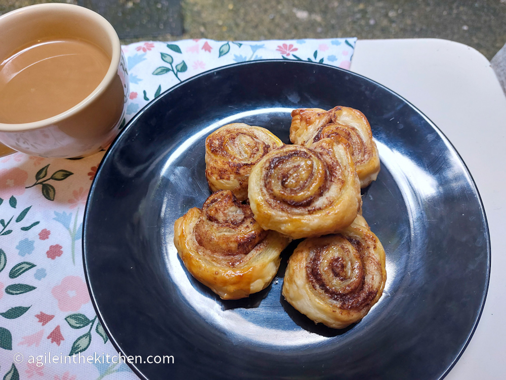 Cinnamon swirls laid on top of each other on a black plate, next to a cup of coffee with milk, all on a background of flowery tablecloth.