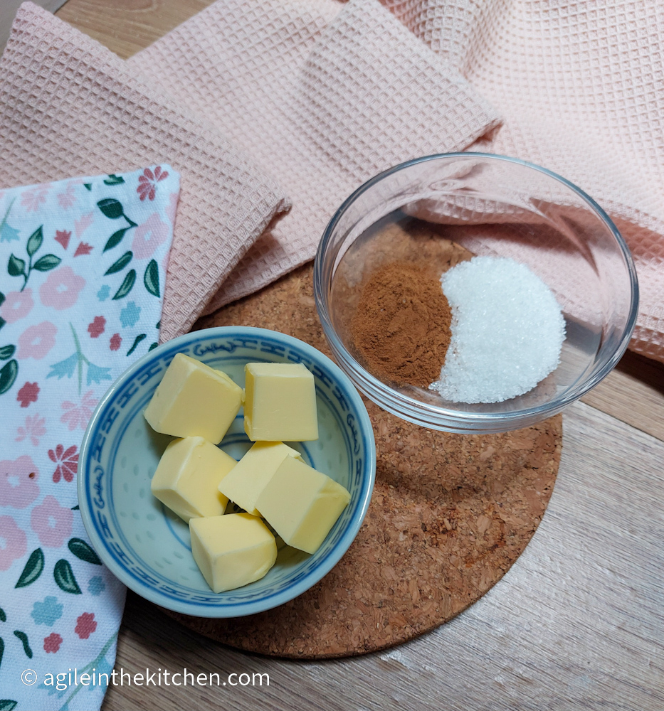 Ingredients for the cinnamon swirl filling, diced butter in a blue bowl and sugar and cinnamon in a glass bowl. In the background a pink fabric and a flowery table cloth