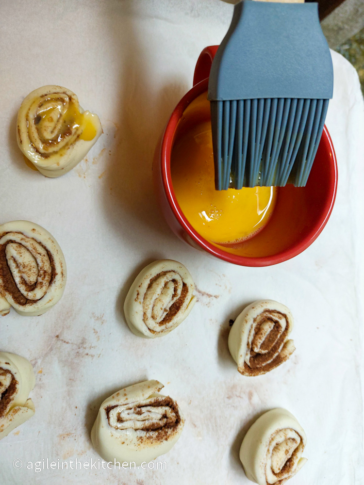 Cinnamon swirls rolled up and cut into smaller pieces, laying cut side up, ready to be brushed with egg wash in a red cup.