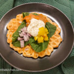 On a green background, a black plate with a waffle topped with fresh spinach, red onion, yellow peppers, smoked salmon, creamy wasabi sauce, and a sprinkle of white sesame seeds