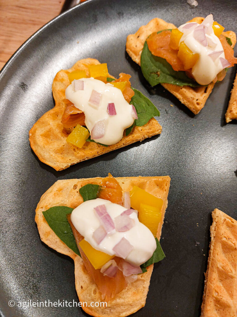 Waffles cut in segments to resemble hearts, topped with a fresh spinach leaf, smoked salmon, diced red onions and yellow peppers, and a dollop of creamy wasabi dressing