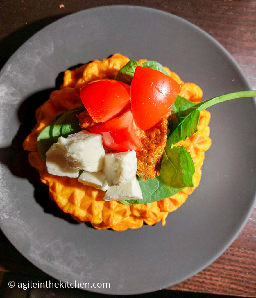 Waffle on a grey plate topped with fresh spinach, chopped tomatoes and mozzarella, and red pesto