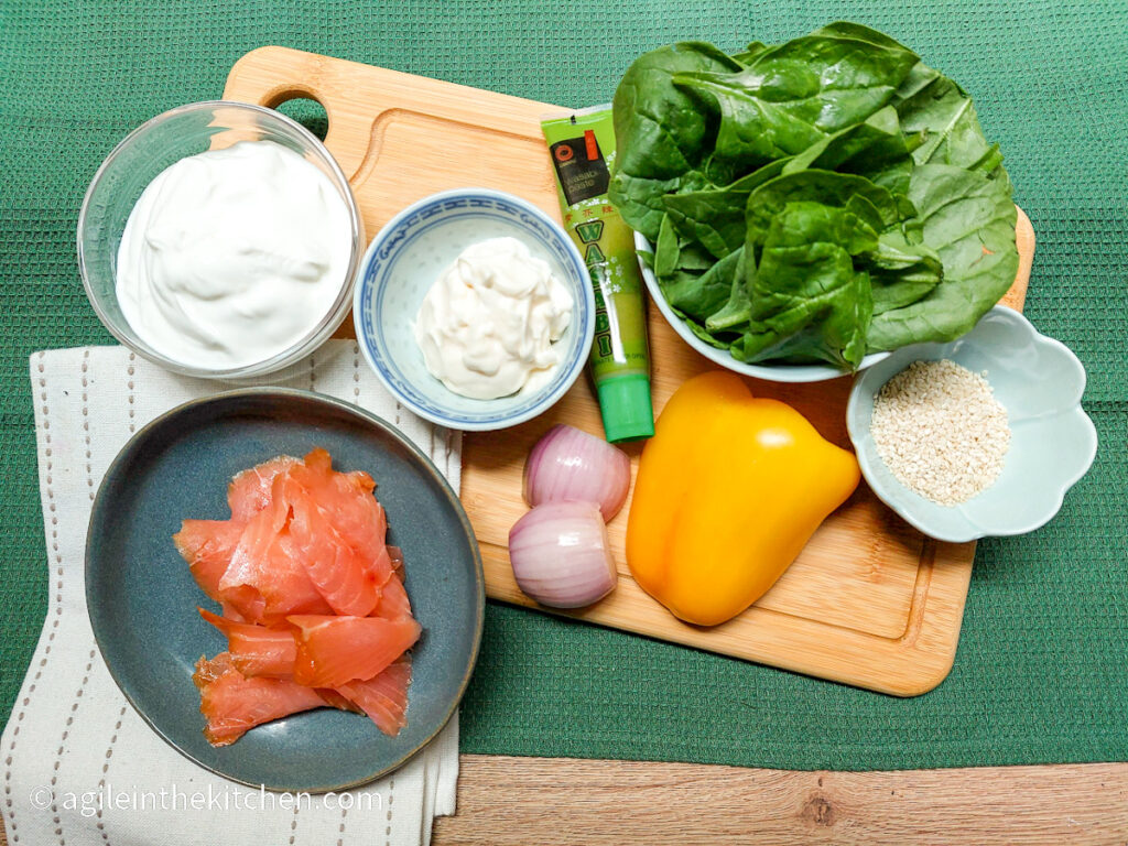 Ingredients to add to waffles laid out on a wooden chopping board, smoked salmon on a blue plate, Greek yogurt, mayonnaise, a red onion cut in half, wasabi in a tube, washed fresh spinach, half of a yellow pepper, white sesame seeds