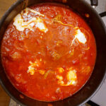 Shakshuka cooking in a large sauteuse, showing traces of poached eggs poking out of the red sauce.