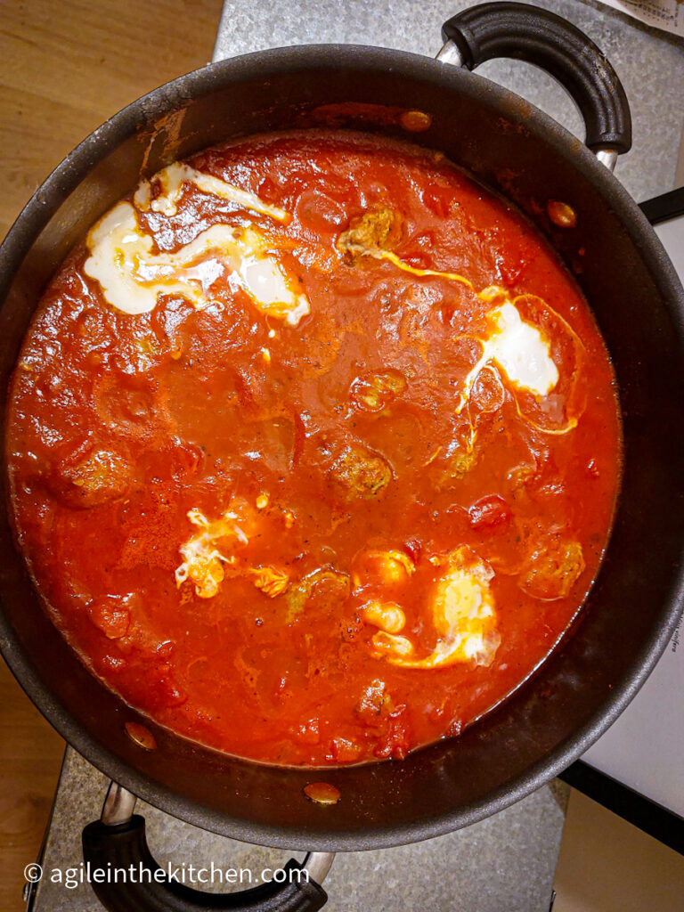 Shakshuka cooking in a large sauteuse, showing traces of poached eggs poking out of the red sauce.