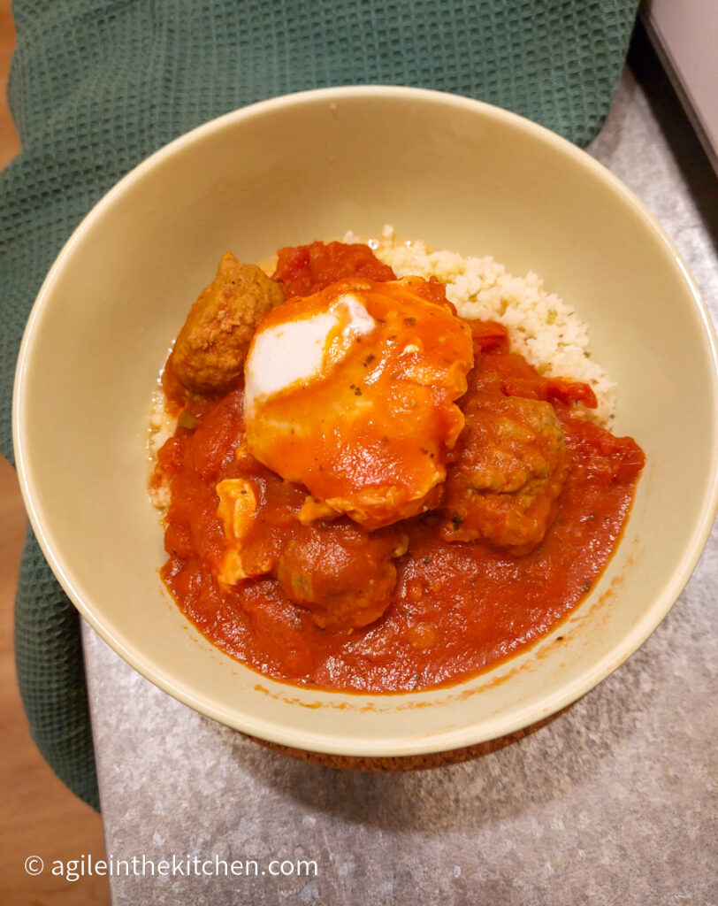 Shakshuka which is Moroccan meatballs in a red sauce and a poached egg is plated in a green bowl on top of couscous. Next to a green cloth.