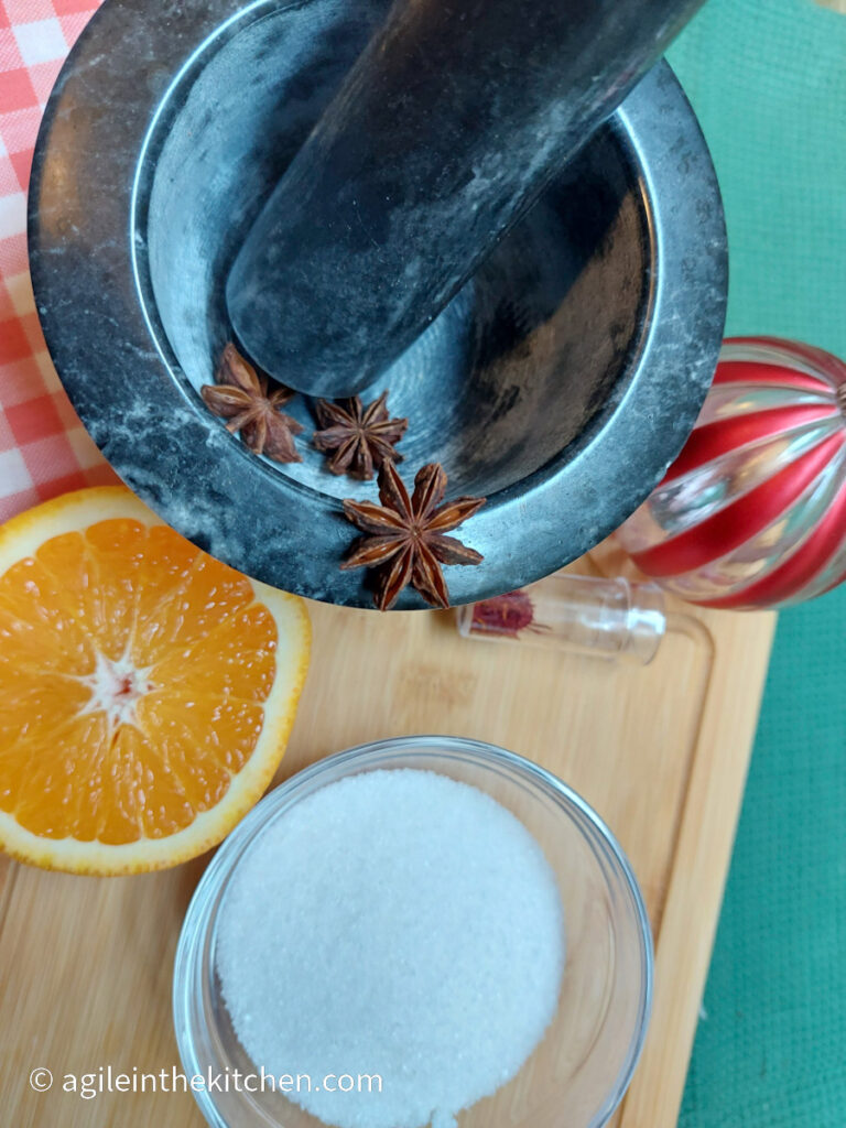 Ingredients to make saffron candied orange slices with star anise. Three star anise, a pinch of saffron strings, a glass bowl of sugar and half of an orange. In the background, resting on a wooden cutting board, a stone mortar and pestle, a red striped Christmas bauble and a red gingham cloth.