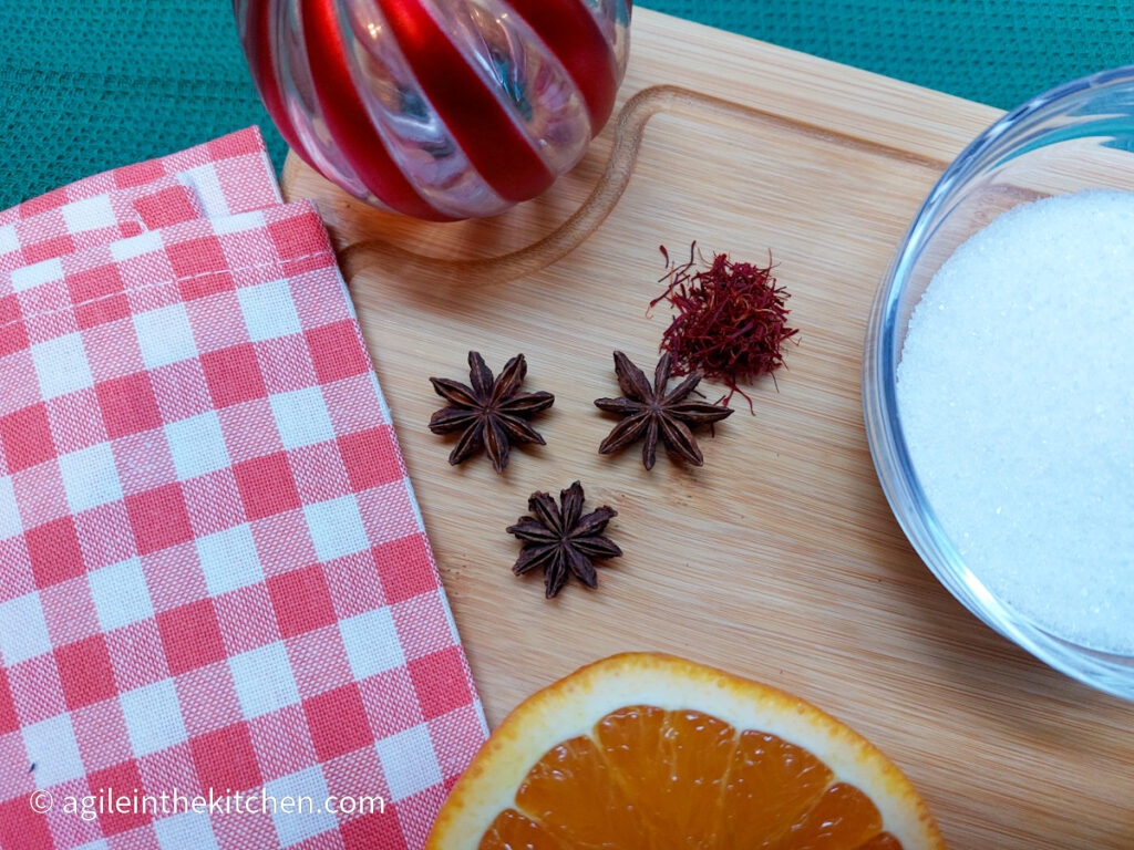 Ingredients to make saffron candied orange slices with star anise. Three star anise, a pinch of saffron strings, a glass bowl of sugar and half of an orange. In the background a wooden cutting board, a red striped Christmas bauble and a red gingham cloth.