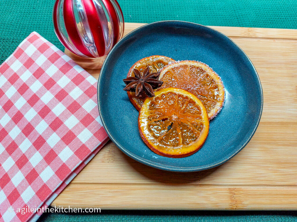 Three saffron candied orange slices are laid out on a blue plate with a decorative star anise. In the background is a red striped Christmas bauble and a red gingham cloth.