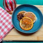 Three saffron candied orange slices are laid out on a blue plate with a decorative star anise. In the background is a red striped Christmas bauble and a red gingham cloth.