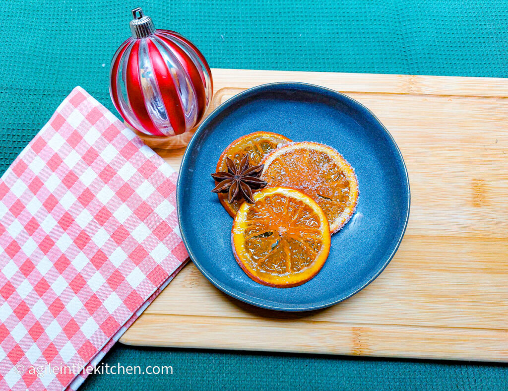 Three saffron candied orange slices are laid out on a blue plate with a decorative star anise. In the background is a red striped Christmas bauble and a red gingham cloth.