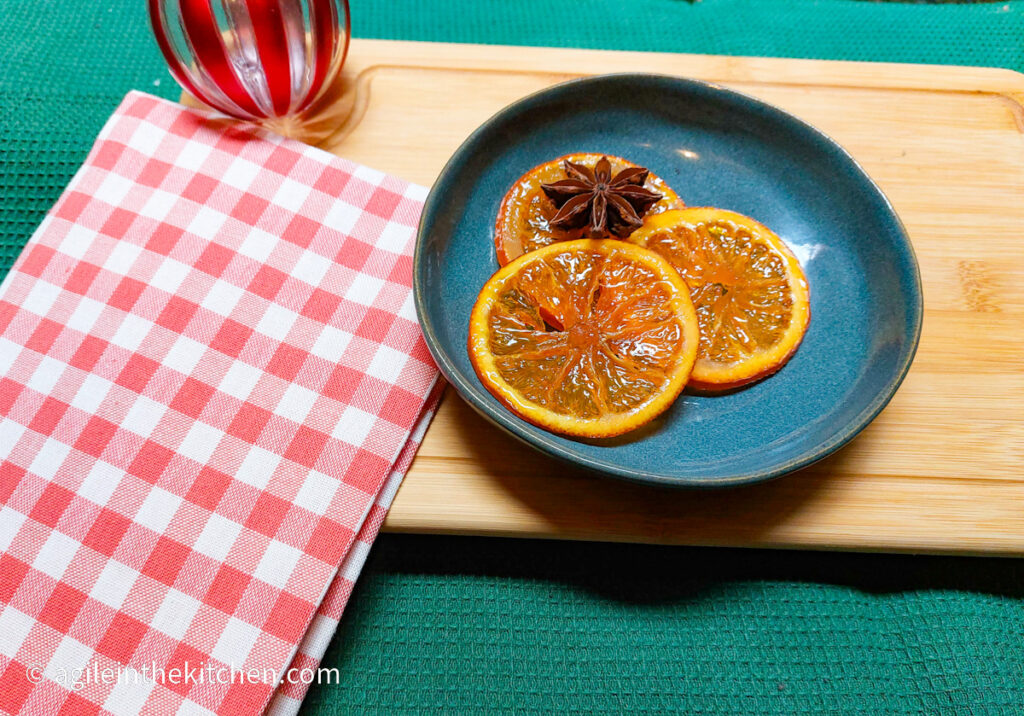 Three saffron candied orange slices are laid out on a blue plate with a decorative star anise. In the background is a red striped Christmas bauble and a red gingham cloth.