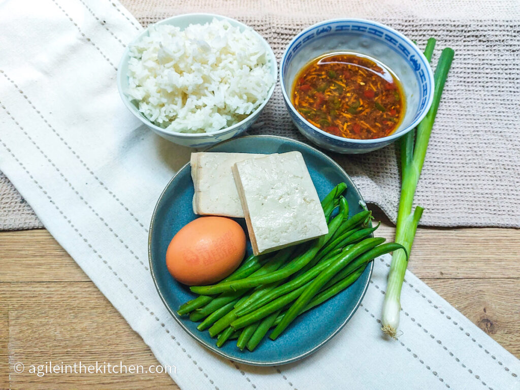 Everything stir fry ingredients laid out on two beige cloths. Cooked rice in a bowl, stir fry sauce mixed in a bowl, a blue plate with an egg, two slices of tofu and some green beans, a stalk of green onion.