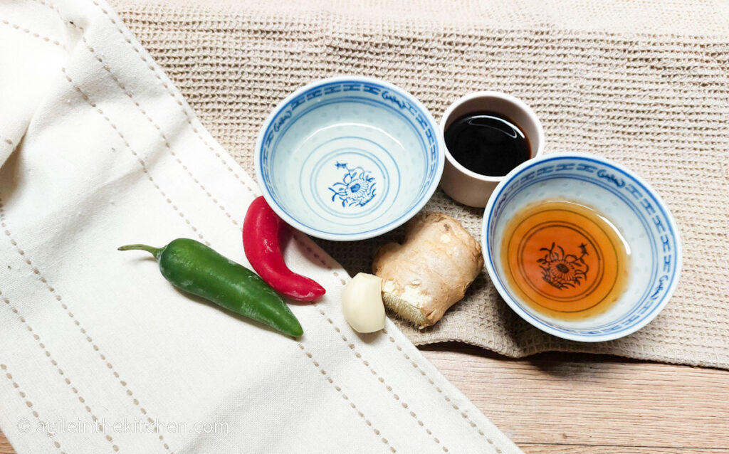 Everything stir fry sauce ingredients laid out: two small chilies one green and one red, a bowl of white wine vinegar, a clove of garlic, a thumb size piece of ginger a bowl of soy sauce and a bowl of sesame oil. All on a background of two beige cloths.