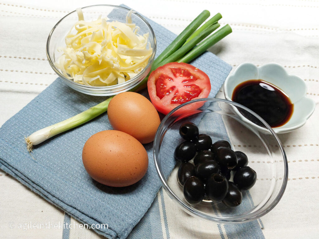 Ingredients to an everything omelet is laid out on a blue cloth. Shredded cheese in a glass bowl, a stalk of green onion, two eggs, half a tomato, a bunch of black olives in a glass bowl and a bowl of balsamic vinegar.