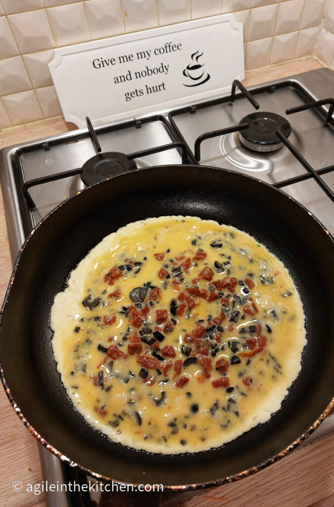 An everything omelet is cooking in a black frying pan on a gas hob.