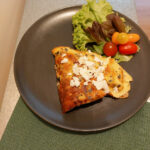 Everything omelet plated on a black plate on top of a green cloth, the omelet is folded in quarters and sprinkled with feta cheese. There is also a green salad with some tomatoes on the same plate.