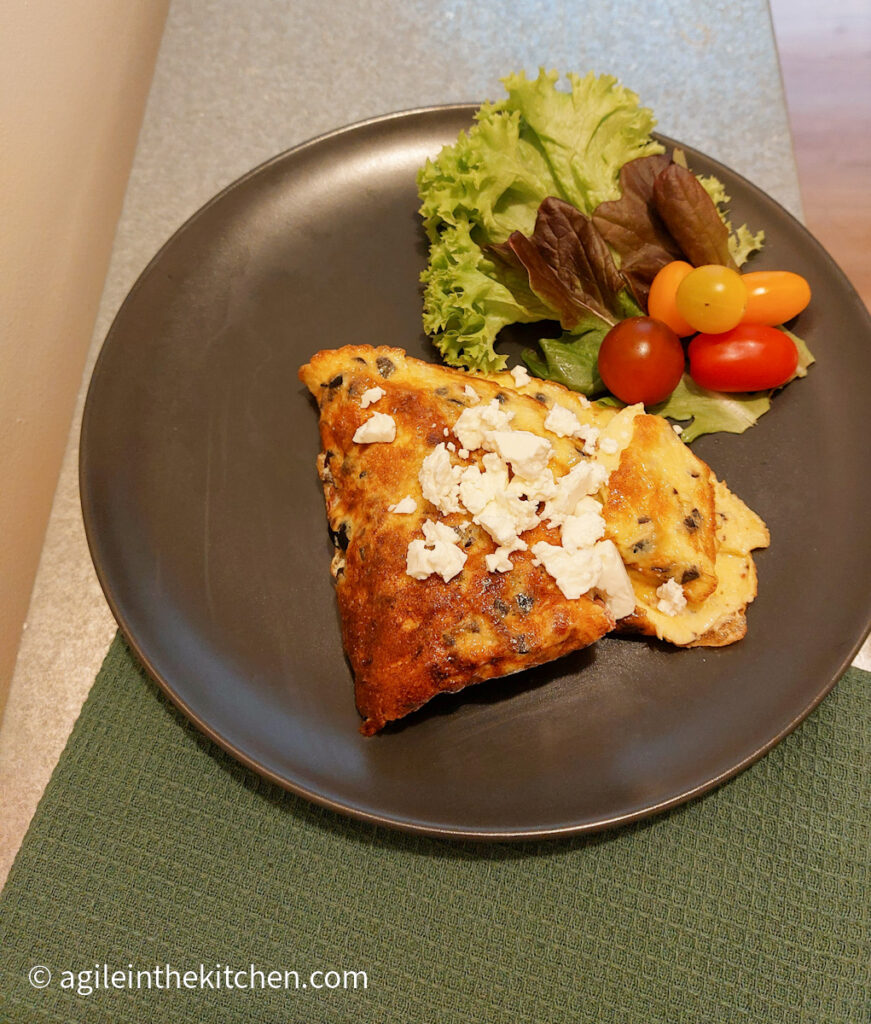 Everything omelet plated on a black plate on top of a green cloth, the omelet is folded in quarters and sprinkled with feta cheese. There is also a green salad with some tomatoes on the same plate.