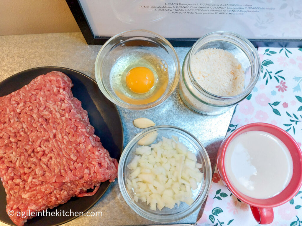 Ingredients for traditional Swedish meatballs lined up; mincemeat on a black plate, an egg cracked in a bowl, breadcrumbs, two cloves of garlic, onions cut into smaller pieces, a cup of milk