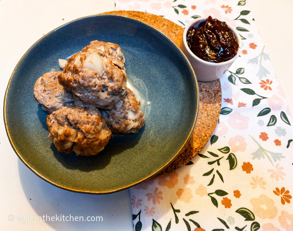 Four baked meatballs piled up on a blue plate with a pot of lingonberry jam next to it.