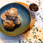 Four baked meatballs piled up on a blue plate with a pot of lingonberry jam next to it.