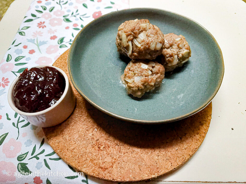 Four baked meatballs piled up on a blue plate with a pot of lingonberry jam next to it.