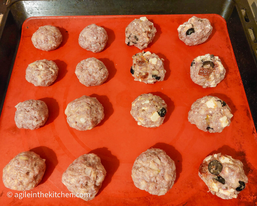 Sixteen uncooked meatballs lined up on a cooking sheet on a red silicone baking sheet. Some are plain and some are with added olives, feta cheese and sun dried tomatoes.