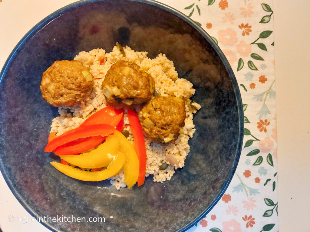 Finished Moroccan meatballs plated in a blue bowl on top of couscous and next to sliced red and yellow bell peppers.