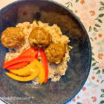 Finished Moroccan meatballs plated in a blue bowl on top of couscous and next to sliced red and yellow bell peppers.