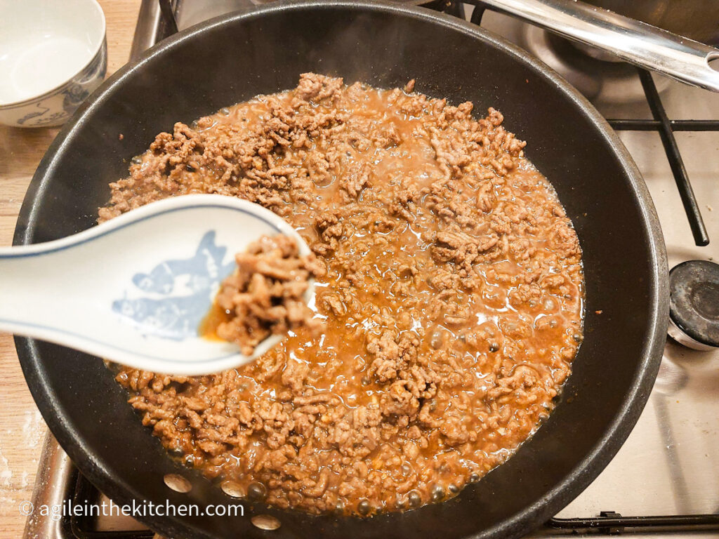 Mince meat in a frying pan cooked on a gas stove top with added Asian sauce mix, a large porcelain spoon in the foreground with some mince.