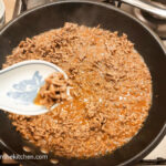 Mince meat in a frying pan cooked on a gas stove top with added Asian sauce mix, a large porcelain spoon in the foreground with some mince.