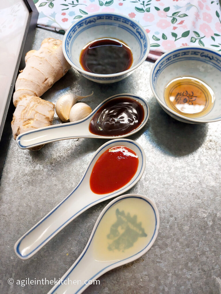 Ingredients to Asian sauce in different containers. On three large spoons lime juice, shiraza, hoisin. In two bowls soy sauce, sesame oil. On the bench two cloves of garlic, on stump of fresh ginger