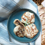 Three Asian mince buns on a blue plate, one cut open to expose the filling, with a few green onions on top, laying in a weaved basket with a blue and white stripped towel.