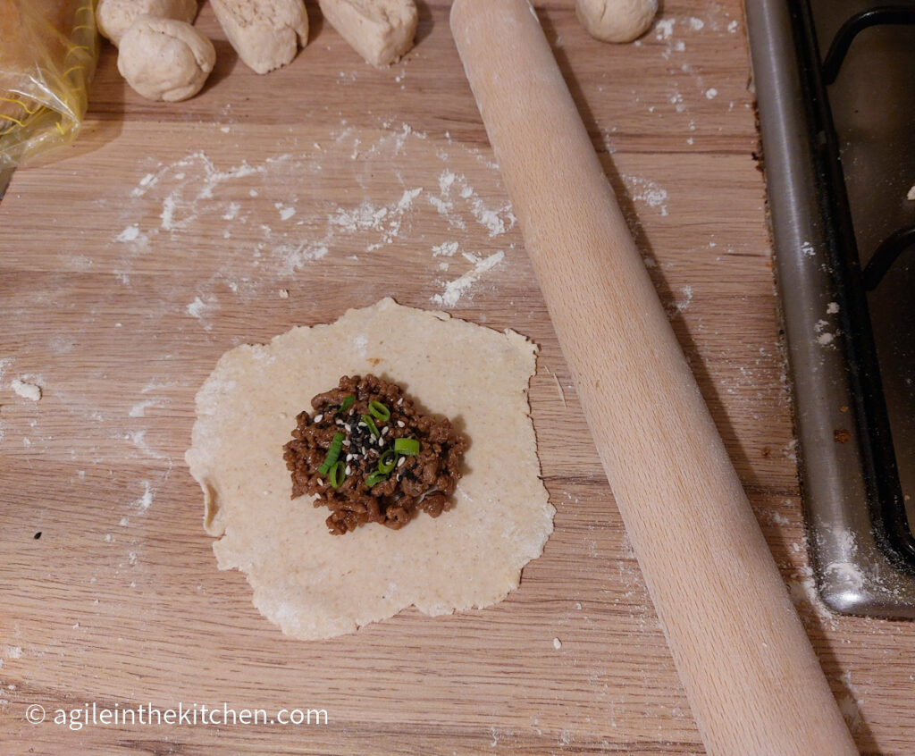A flatbread rolled out on a wooden counter top, topped with Asian mince, chopped green onions and a sprinkle of sesame seeds, next to a rolling pin. At the top of the picture, five dough balls waiting to be rolled out.