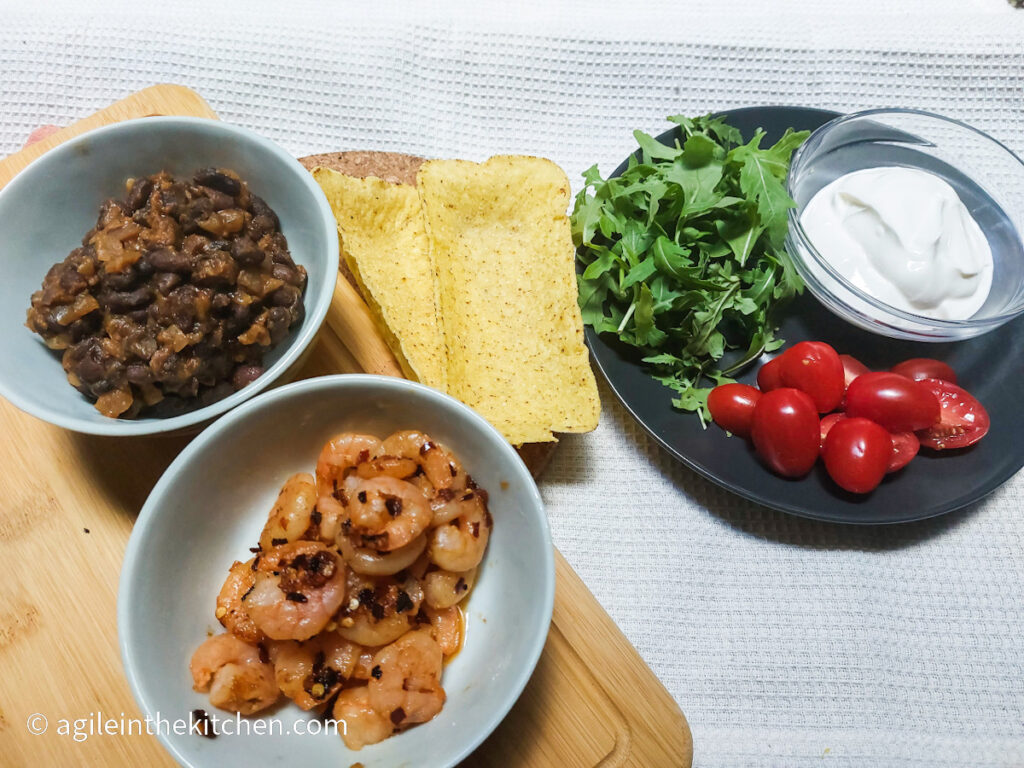 Ingredients to put together a black bean shrimp taco, cooked black beans in a bowl, cooked spicy shrimp in a bowl, two mini taco tubs, arugula, cherry tomatoes and creme fraiche on a plate.