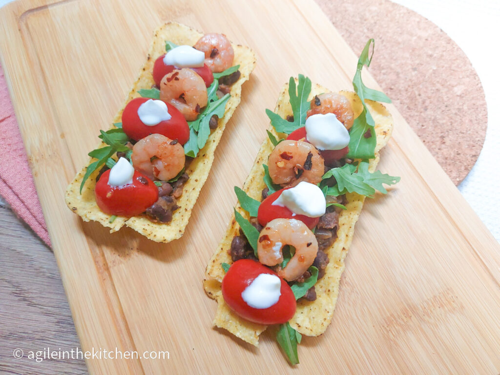 A wooden chopping board with two mini taco tubs filled with black beans, arugula, red cherry tomatoes, spicy shrimp and dollops of creme fraiche.