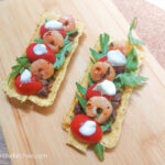 A wooden chopping board with two mini taco tubs filled with black beans, arugula, red cherry tomatoes, spicy shrimp and dollops of creme fraiche.