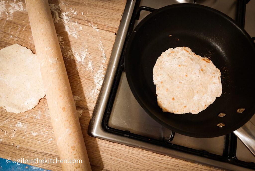 Flatbread baking in a black frying pan on a gas stove top. To the left one flatbread is being rolled out, using a wooden rolling pin.
